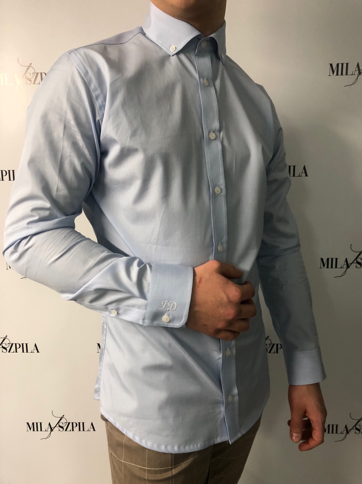 Jasnoniebieska koszula z długim rękawem i monogramem 'JD' na mankiecie, zapinana na białe guziki, widoczna na tle z logotypem.