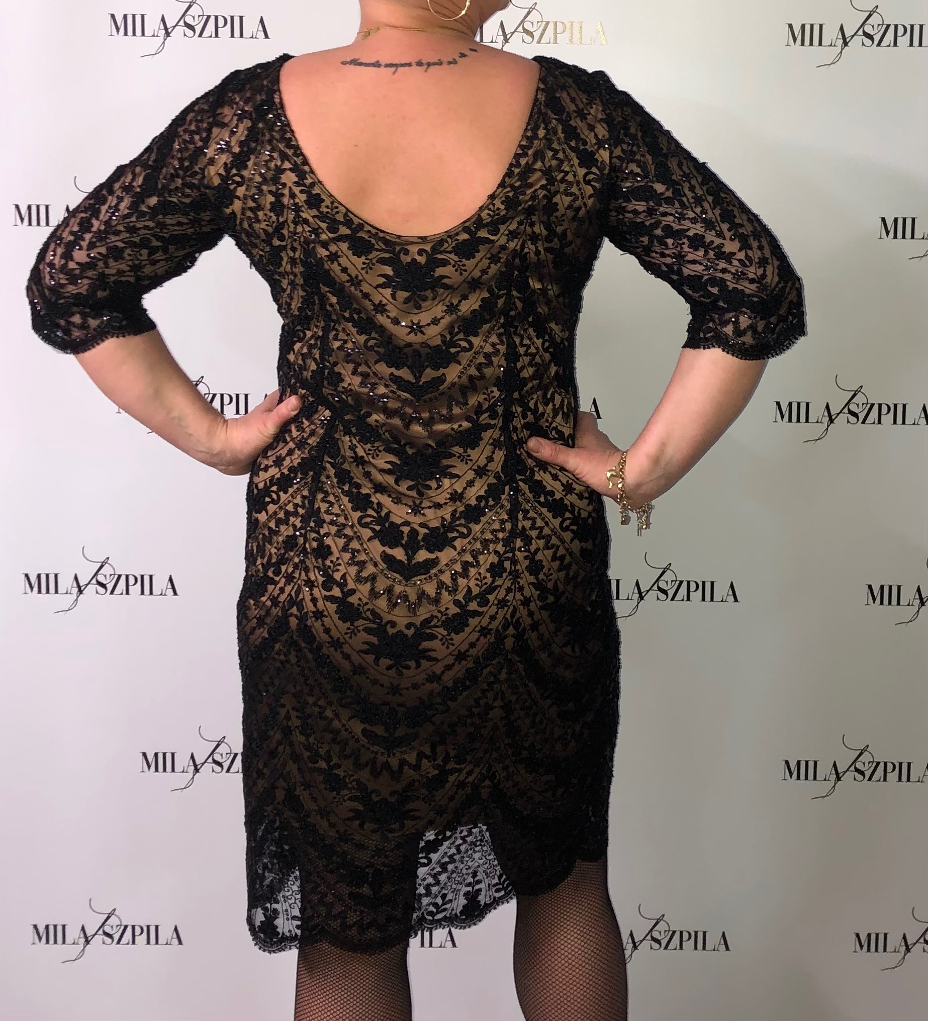 Czarna, koronkowa sukienka z krótkim rękawem, widziana z tyłu na kobiecie w rajstopach, na tle ściany z powtarzającym się logo 'MILA SZPILA'.