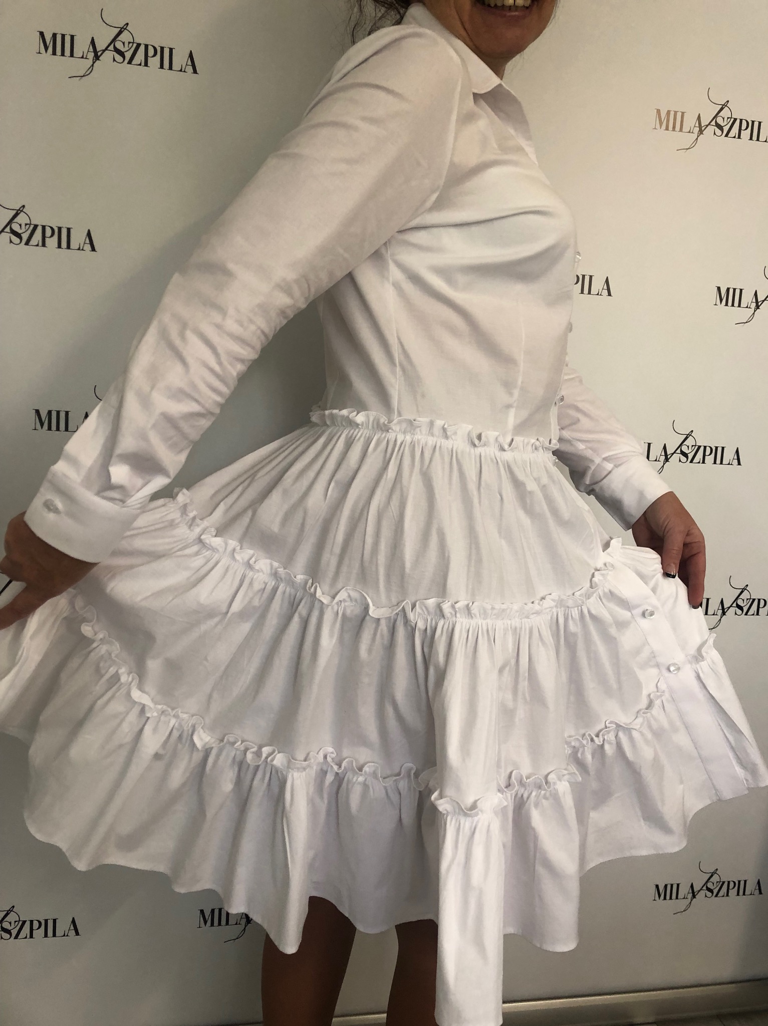 Biała, rozkloszowana sukienka z falbanami, połączenie koszuli z dołem z kilku warstw falban, prezentowana na osobie na tle ściany z logo.