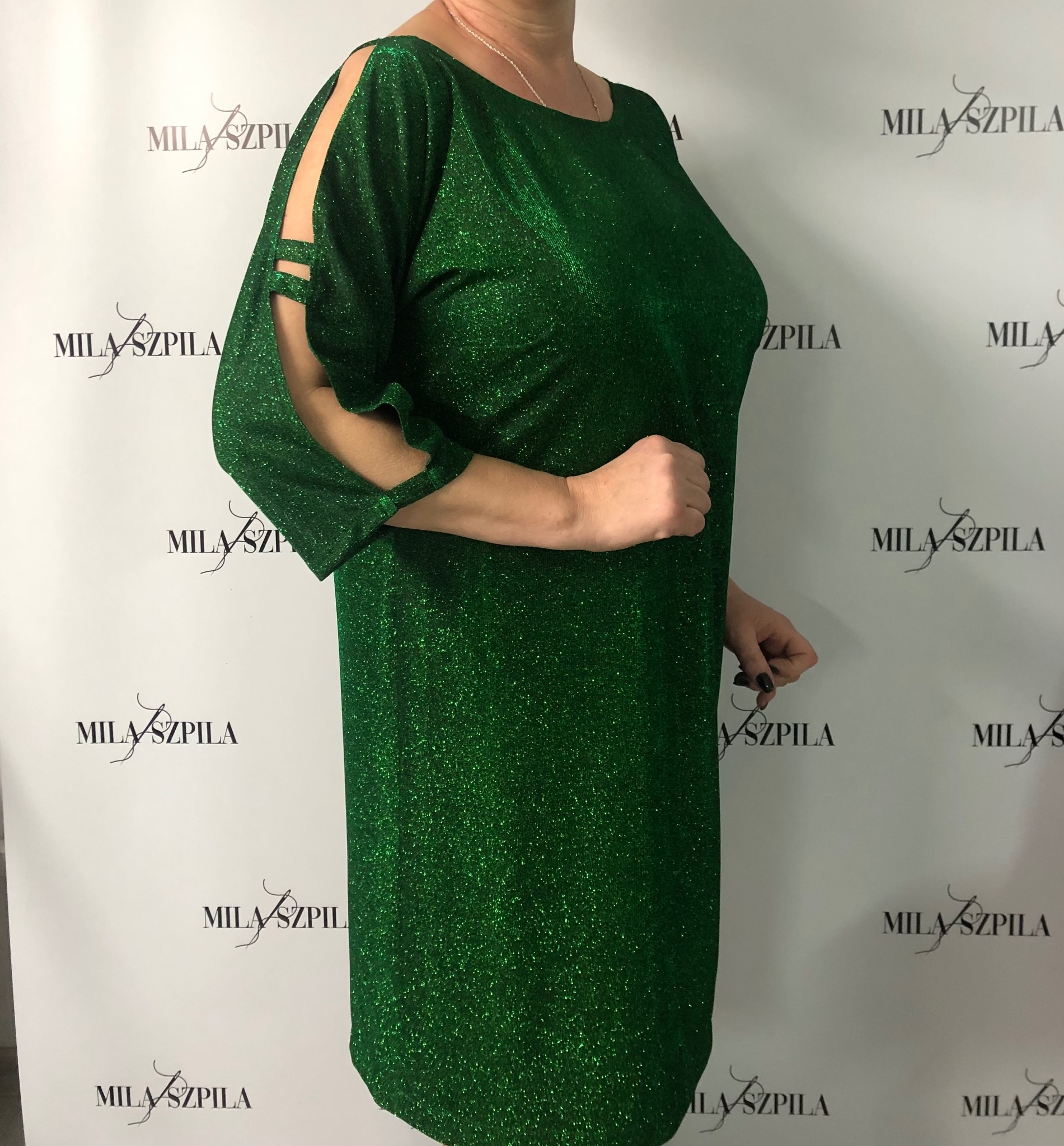 Błyszcząca, szmaragdowa sukienka mini z rozcięciami na ramionach, prezentowana na tle z powtarzającym się logo 'MILA SZPILA'.