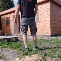 Paweł Stasierowski - Mężczyzna w koszulce z logo firmy stoi przed nowo wybudowanym, drewnianym domkiem z oknem i drzwiami, widoczne drewniane palety i ślady po budowie.