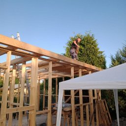 Paweł Stasierowski - Pracownik w koszulce na drewnianej konstrukcji budynku w trakcie budowy, widoczne narzędzia i drabina, w tle zielone drzewa i błękitne niebo.