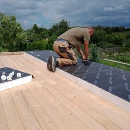 Paweł Stasierowski - Pracownik na kolanach mocuje membranę dachową do drewnianej konstrukcji dachu domu jednorodzinnego, widok na ogród w tle.