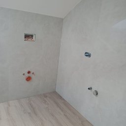 Paweł Stasierowski - Surowa łazienka w trakcie wykańczania: szare płytki imitujące kamień na ścianach, drewnopodobna podłoga, widoczne instalacje hydrauliczne i elektryczne przed montażem armatury.