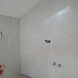 Paweł Stasierowski - Świeżo wykafelkowana łazienka z drewnopodobną podłogą, widoczne przygotowane punkty instalacyjne na ścianie i wystające rury kanalizacyjne.