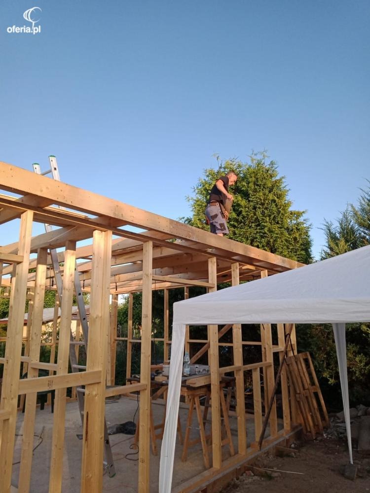 Pracownik w koszulce na drewnianej konstrukcji budynku w trakcie budowy, widoczne narzędzia i drabina, w tle zielone drzewa i błękitne niebo.
