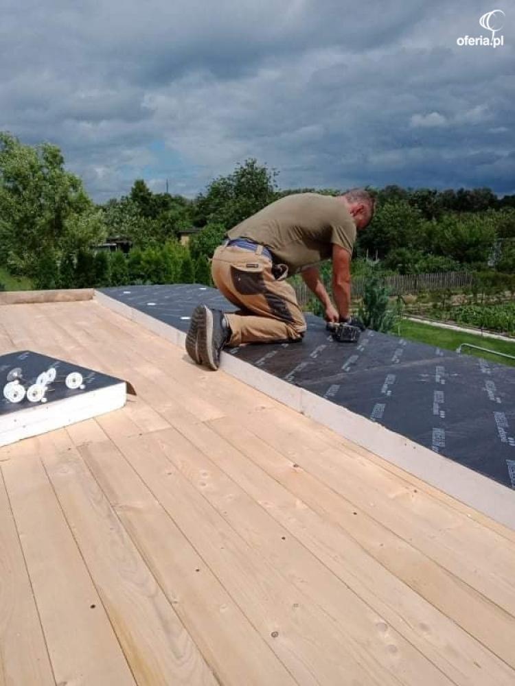 Pracownik na kolanach mocuje membranę dachową do drewnianej konstrukcji dachu domu jednorodzinnego, widok na ogród w tle.