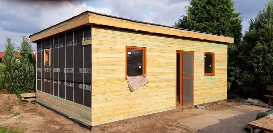 Drewniany budynek gospodarczy z oknami i drzwiami, częściowo ocieplony czarną folią izolacyjną Foltech, widok z zewnątrz na tle zieleni.