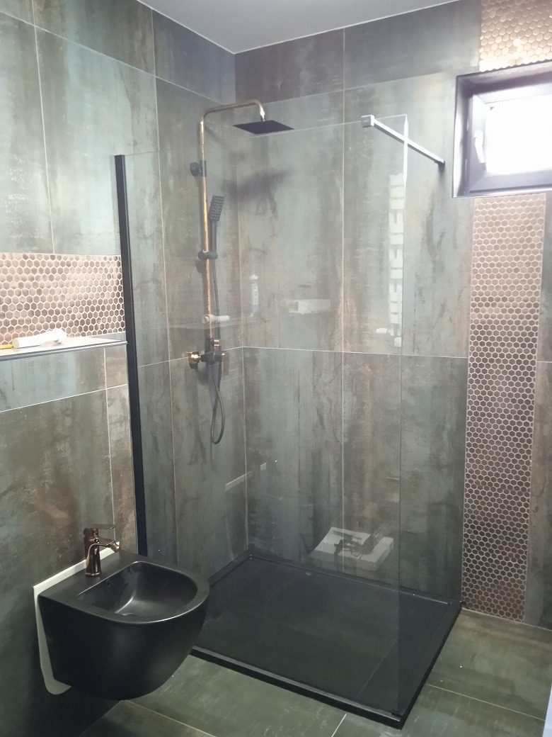 Nowoczesna łazienka z czarną armaturą, kabiną prysznicową typu walk-in z czarnym brodzikiem i podwieszanym bidetem, wykończona szarą płytką imitującą beton i mozaiką heksagonalną.