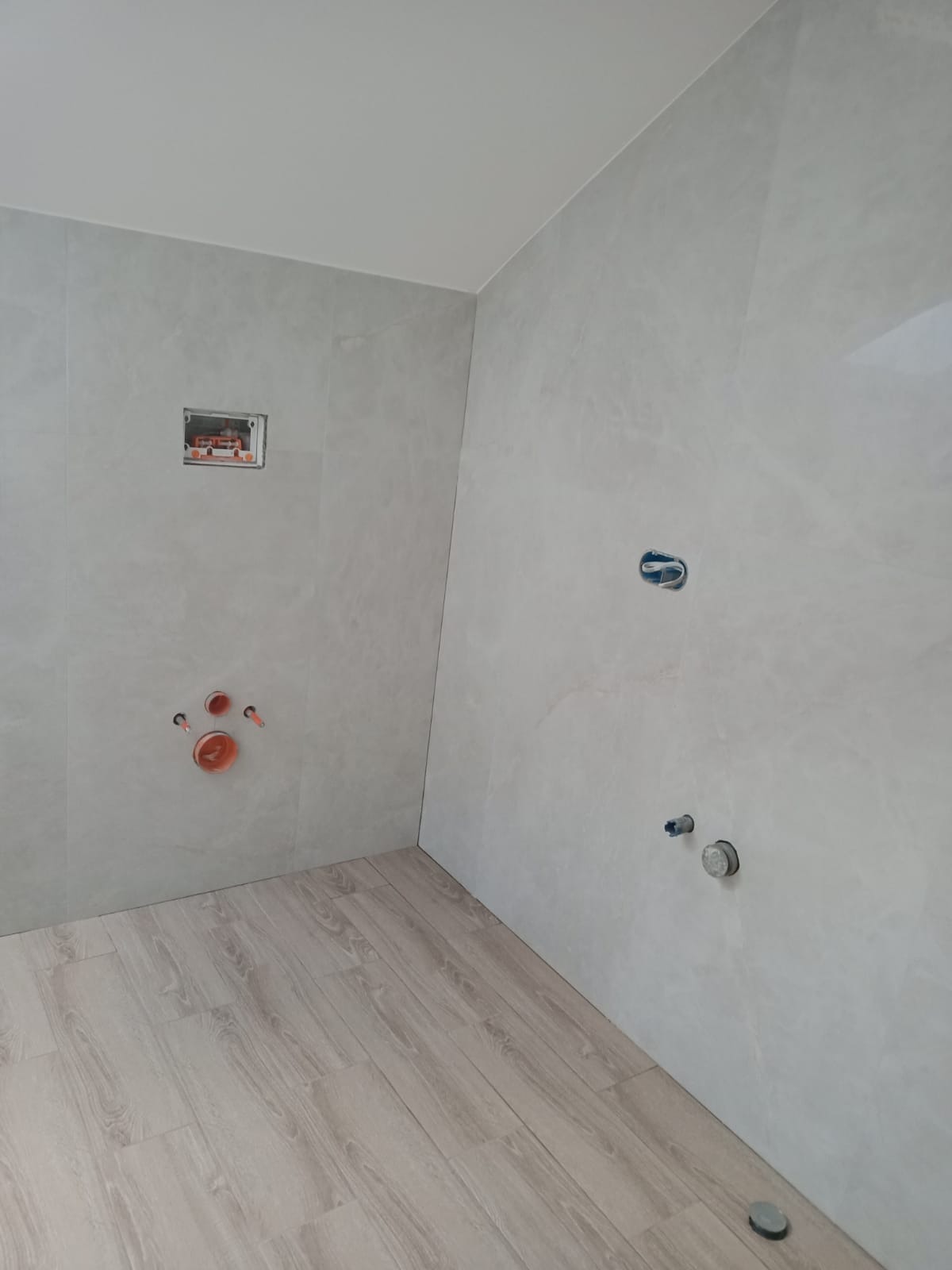 Surowa łazienka w trakcie wykańczania: szare płytki imitujące kamień na ścianach, drewnopodobna podłoga, widoczne instalacje hydrauliczne i elektryczne przed montażem armatury.