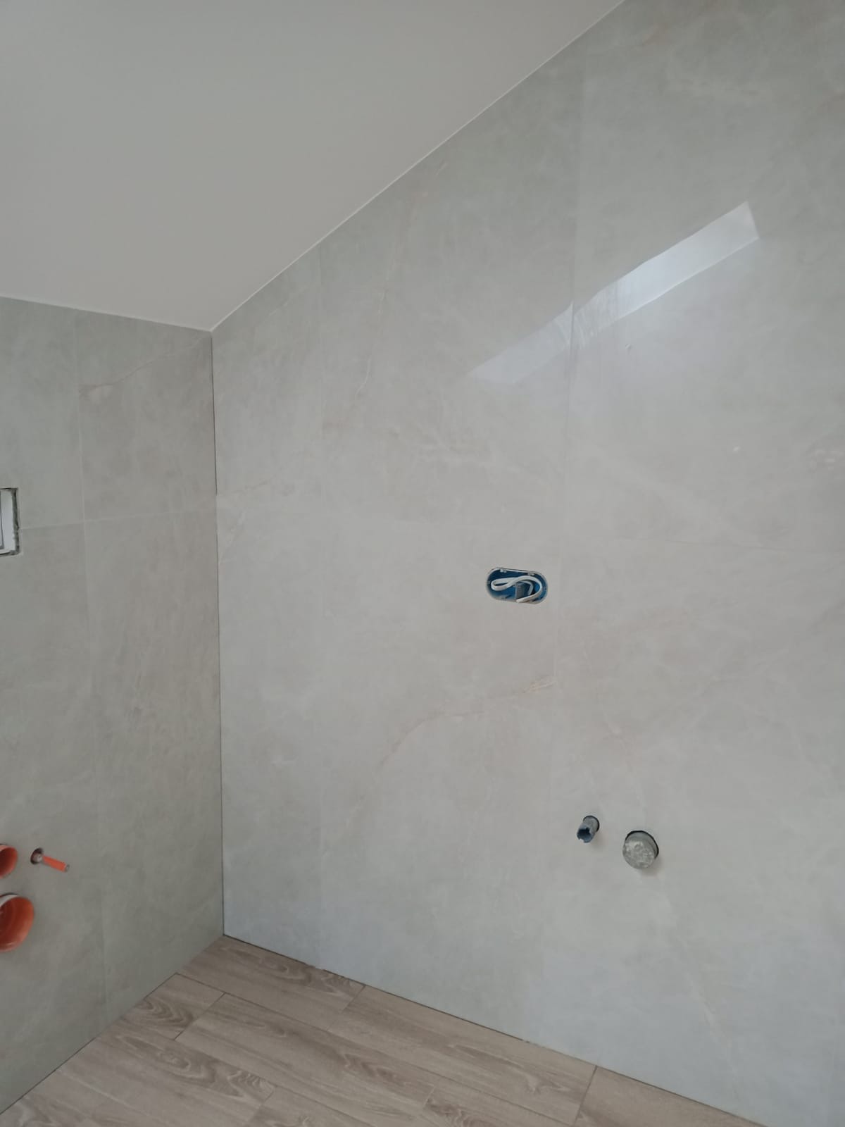 Świeżo wykafelkowana łazienka z drewnopodobną podłogą, widoczne przygotowane punkty instalacyjne na ścianie i wystające rury kanalizacyjne.