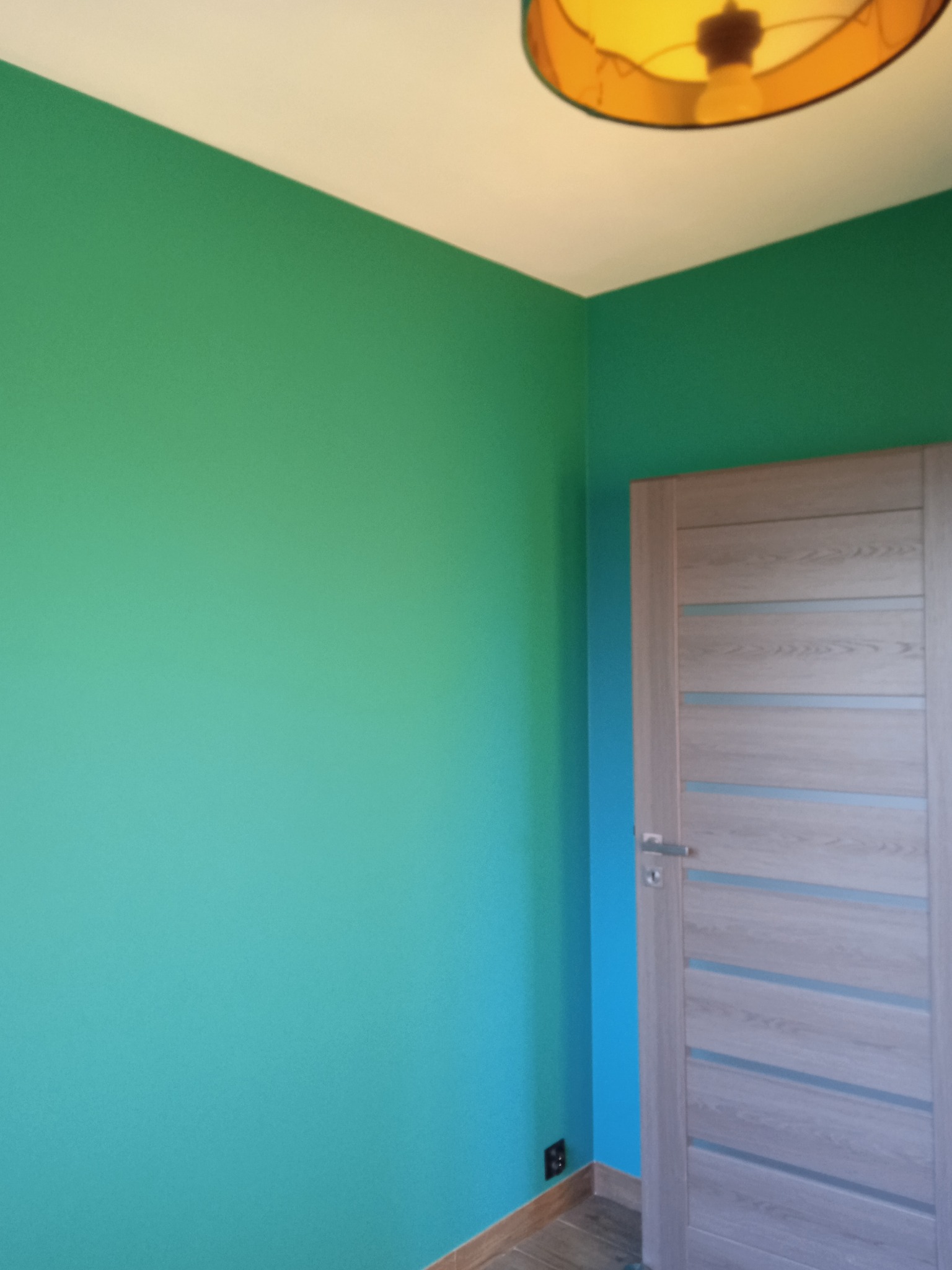 Pomalowane na zielono ściany, jasne drewniane drzwi z poziomymi przeszkleniami, widoczny fragment podłogi i brązowy klosz lampy sufitowej.