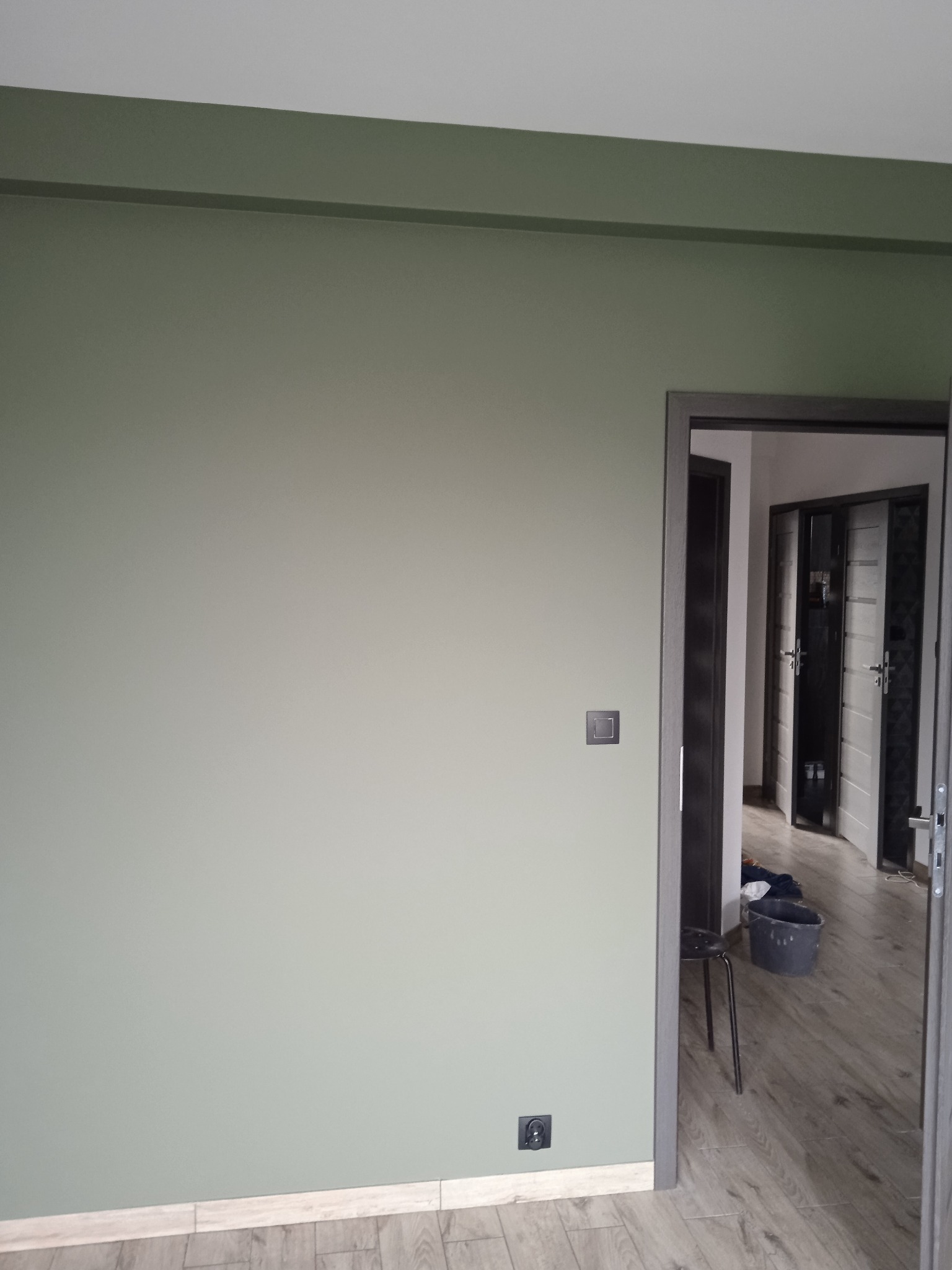 Świeżo pomalowana na oliwkowy kolor ściana z szarą ramą drzwi i widocznym gniazdkiem elektrycznym, z boku widoczne wiadro i krzesło malarskie.