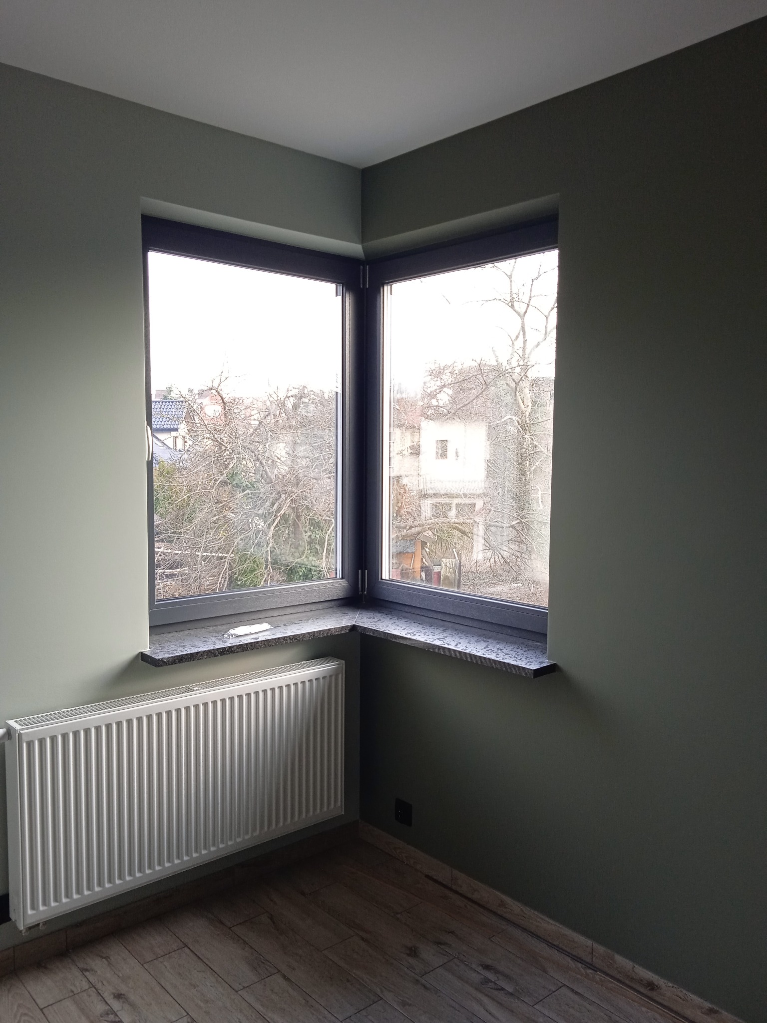 Wnętrze pokoju po remoncie z narożnym oknem w ciemnej ramie, granitowym parapetem, białym grzejnikiem i podłogą imitującą drewno, ściany pomalowane na oliwkowy kolor.