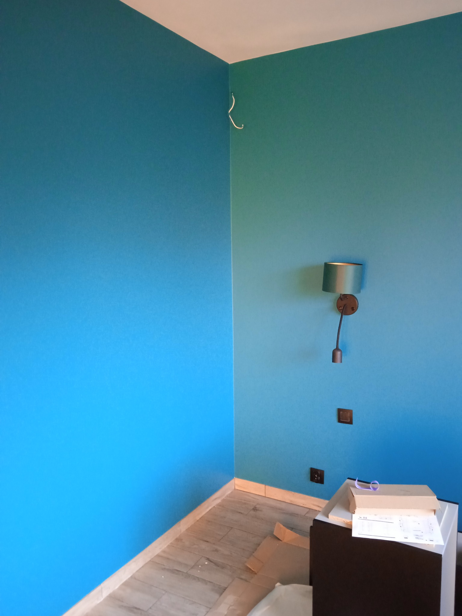 Turkusowe ściany w rogu pokoju, widoczna lampka ścienna z zielonym abażurem, gniazdka elektryczne, listwy przypodłogowe i panele podłogowe, kartony i dokumenty na meblu.