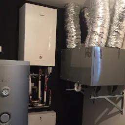 Zainstalowany system grzewczy z szarą obudową, widocznymi rurami i srebrnymi przewodami wentylacyjnymi w pomieszczeniu z czarną ścianą.