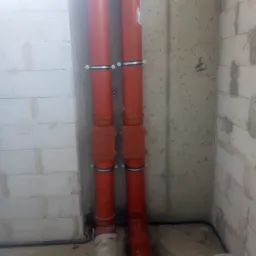 Dwie pionowe rury kanalizacyjne z pomarańczowego PCV, przymocowane metalowymi obejmami do ściany z betonu komórkowego i surowego betonu, widoczne złącza i fragment instalacji elektrycznej w podłodze.