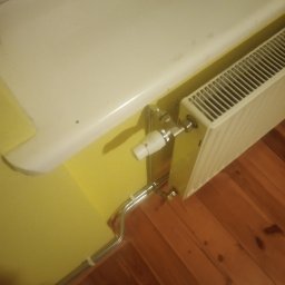 Instalatorstwo sanitarne Jacek Wojciech - Żeberkowy grzejnik pomalowany na żółto zamontowany pod parapetem, z widocznymi rurami doprowadzającymi i termostatem, na tle żółtej ściany i drewnianej podłogi.