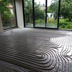 Instalatorstwo sanitarne Jacek Wojciech - Wnętrze w trakcie instalacji ogrzewania podłogowego, widoczne rury grzewcze ułożone na folii z siatką, duże okna z widokiem na zieleń na zewnątrz.