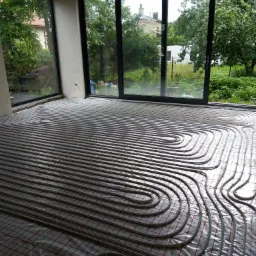 Wnętrze w trakcie instalacji ogrzewania podłogowego, widoczne rury grzewcze ułożone na folii z siatką, duże okna z widokiem na zieleń na zewnątrz.