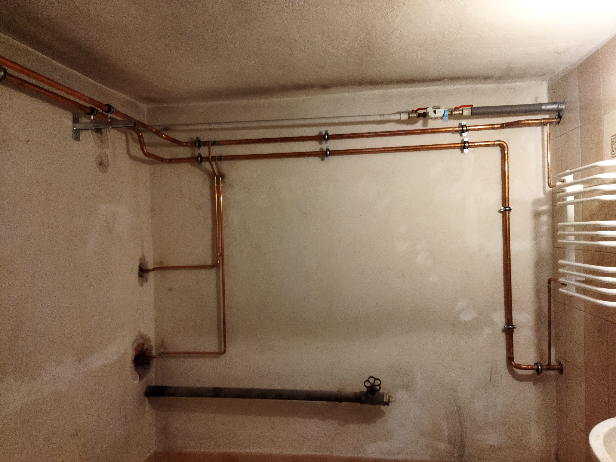 Widok na fragment ściany z zamontowanymi miedzianymi rurami instalacji wodnej i grzewczej, z widocznymi uchwytami mocującymi, zaworem i grzejnikiem drabinkowym w tle.