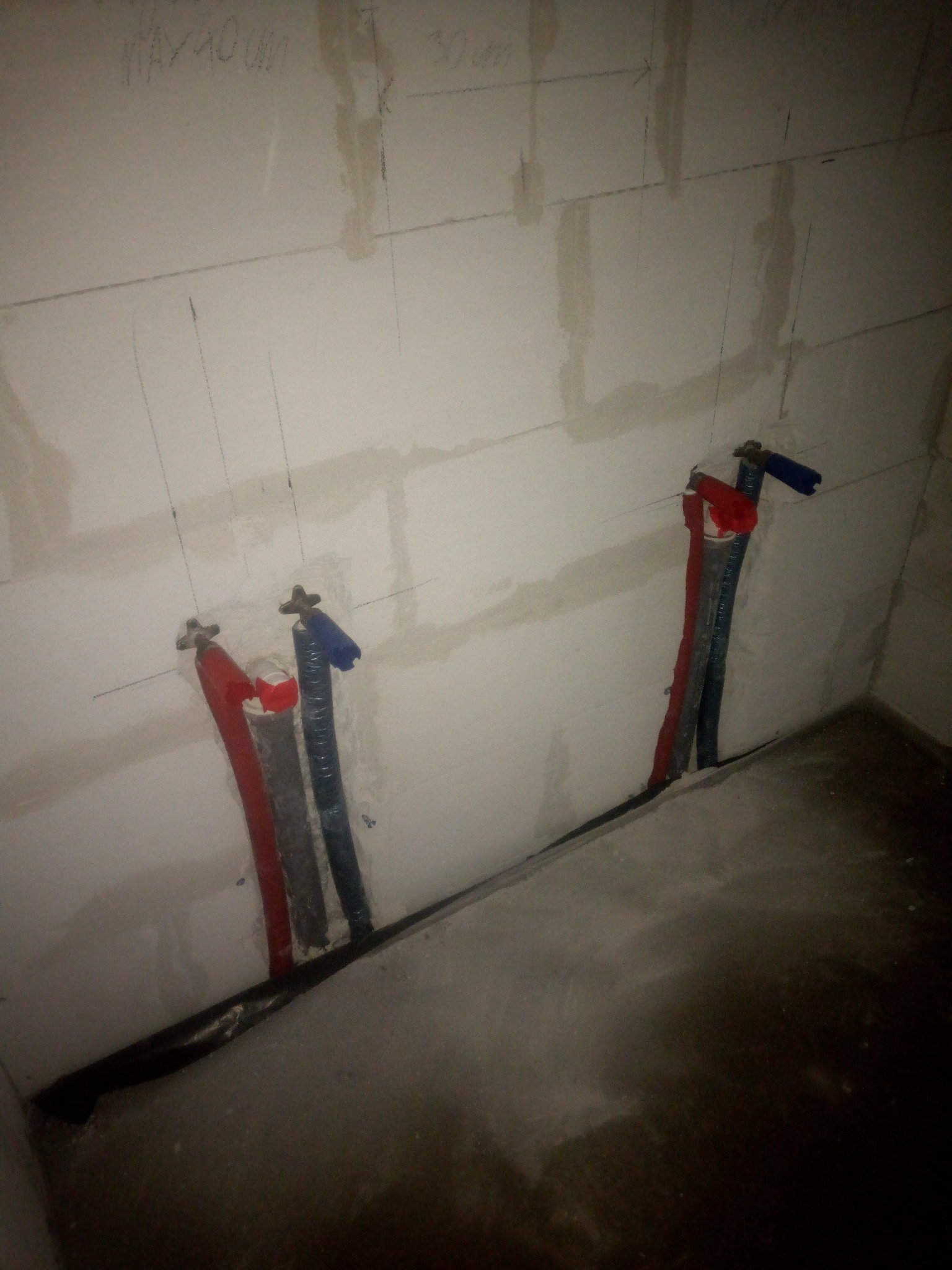 Surowe ściany z wyprowadzonymi rurami instalacji wodnej oznaczonymi kolorami, widoczne linie pomocnicze i wymiary na ścianie.