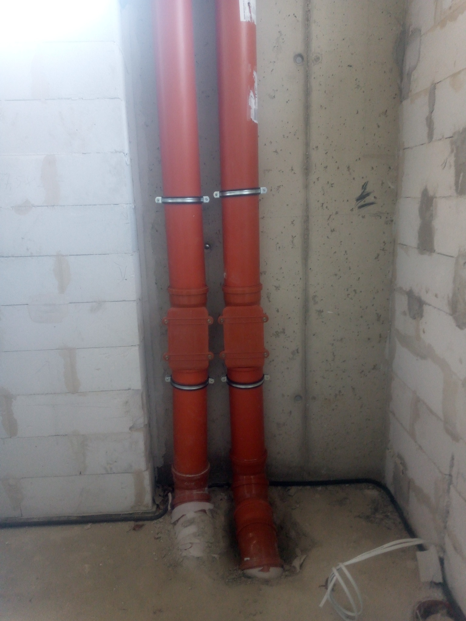 Dwie pionowe rury kanalizacyjne z pomarańczowego PCV, przymocowane metalowymi obejmami do ściany z betonu komórkowego i surowego betonu, widoczne złącza i fragment instalacji elektrycznej w podłodze.