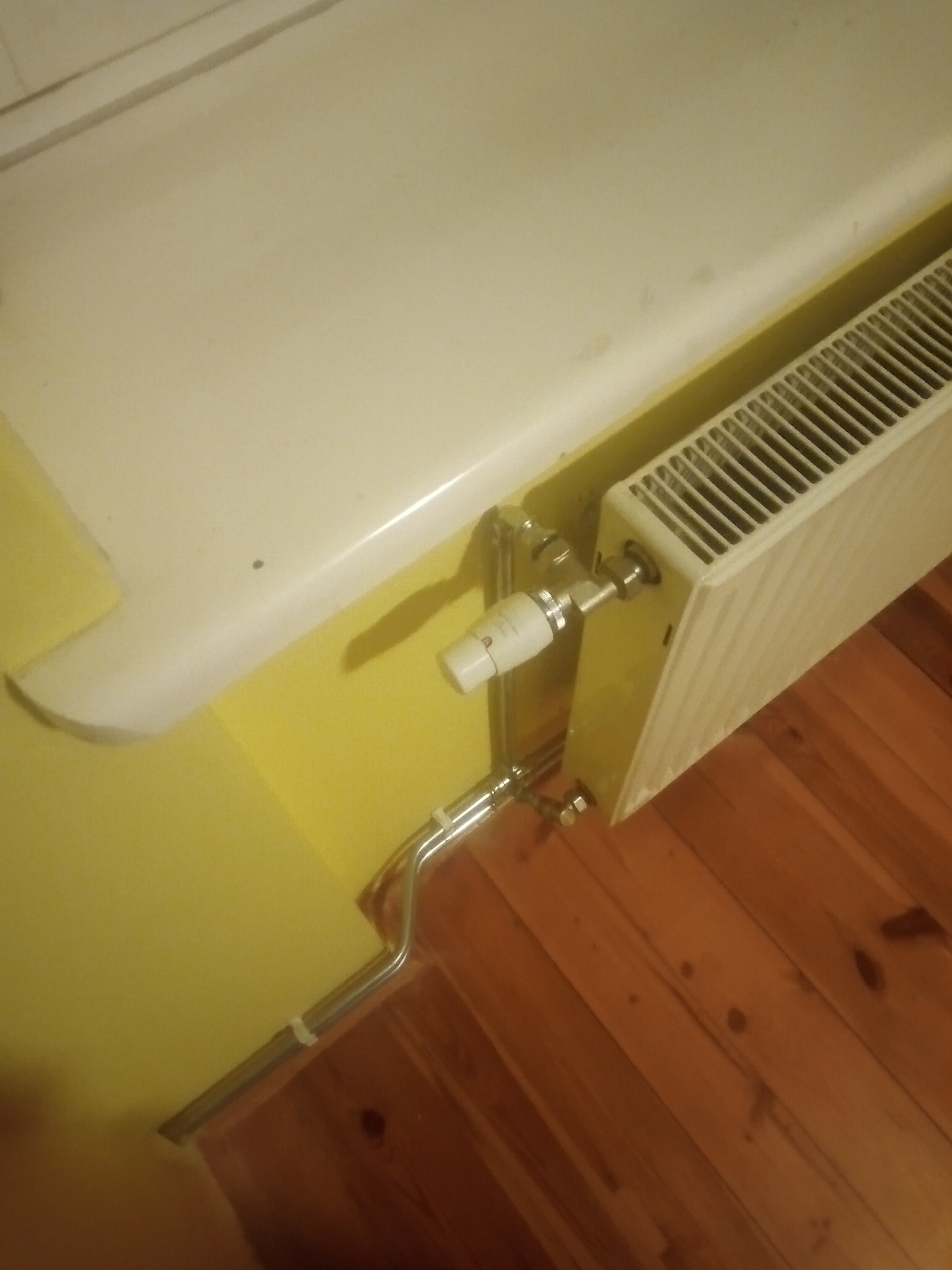 Żeberkowy grzejnik pomalowany na żółto zamontowany pod parapetem, z widocznymi rurami doprowadzającymi i termostatem, na tle żółtej ściany i drewnianej podłogi.