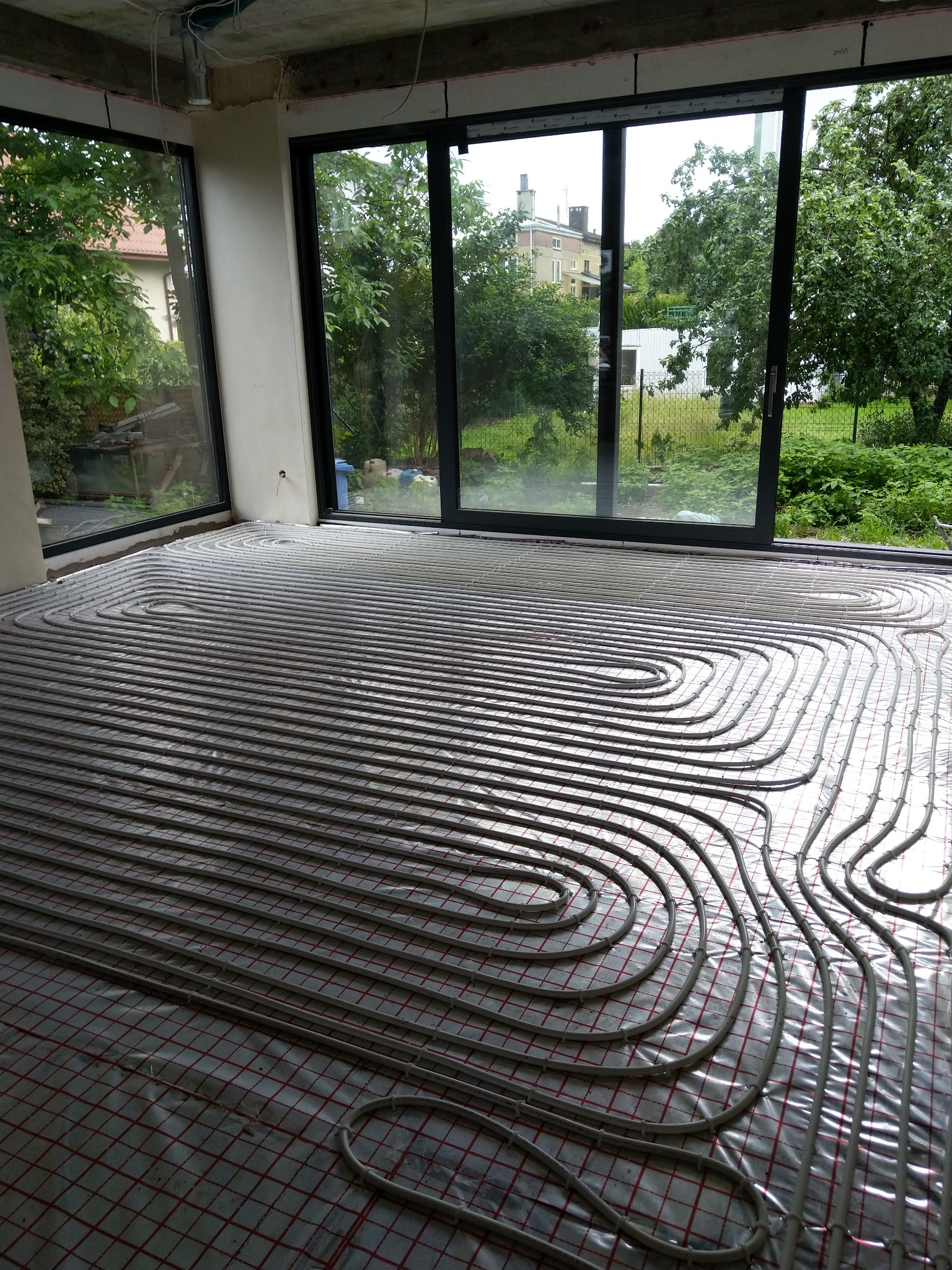 Wnętrze w trakcie instalacji ogrzewania podłogowego, widoczne rury grzewcze ułożone na folii z siatką, duże okna z widokiem na zieleń na zewnątrz.