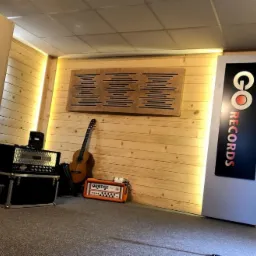 Wnętrze studia z drewnianą boazerią, panelem akustycznym, gitarą akustyczną, wzmacniaczem Orange i sprzętem nagłośnieniowym, obok pionowy szyld z napisem 'GO RECORDS'.