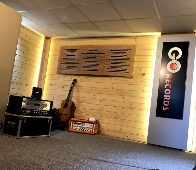 Wnętrze studia z drewnianą boazerią, panelem akustycznym, gitarą akustyczną, wzmacniaczem Orange i sprzętem nagłośnieniowym, obok pionowy szyld z napisem 'GO RECORDS'.