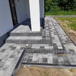 Ułożona kostka brukowa w odcieniach szarości tworząca schody i chodnik przed budynkiem z białą kolumną. Widoczne świeżo ułożone obrzeża i ziemia obok.
