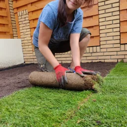Kobieta w czerwonych rękawicach klęczy obok rozwiniętej rolki trawy, z uśmiechem patrzy w obiektyw, w tle drewniane ogrodzenie z ceglanym elementem.