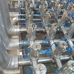 Złożona instalacja przemysłowa z rur i cylindrycznych elementów z podłączonymi niebieskimi kablami, widok z góry na tle kafelkowanej podłogi.