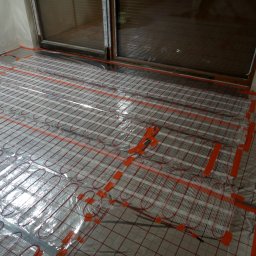 Kama-bud Usługi Ogolnobudowlane Kamil Tylkowski - Instalacja ogrzewania podłogowego: mata grzewcza z czerwonymi przewodami ułożona na podłodze, zabezpieczona pomarańczową taśmą, widoczne fragmenty butów roboczych z odblaskami.