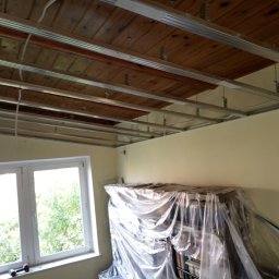 Kama-bud Usługi Ogolnobudowlane Kamil Tylkowski - Montaż metalowej konstrukcji sufitu podwieszanego z płyt kartonowo-gipsowych na tle drewnianego stropu, regał zabezpieczony folią malarską przy oknie.