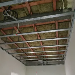 Konstrukcja metalowa podwieszanego sufitu z widoczną izolacją z wełny mineralnej i drewnianymi elementami mocującymi, kable elektryczne zwisające z sufitu.