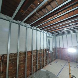 Kama-bud Usługi Ogolnobudowlane Kamil Tylkowski - Stelaż z profili stalowych do płyt karton gips na tle ceglanej ściany i drewnianego stropu z widocznymi kablami, oświetlony lampą budowlaną.