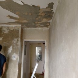 Kama-bud Usługi Ogolnobudowlane Kamil Tylkowski - Pokój w trakcie remontu: uszkodzony sufit z odpadającym tynkiem, ściany przygotowane do prac wykończeniowych, drabina aluminiowa w przejściu do kolejnego pomieszczenia.