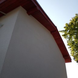Kama-bud Usługi Ogolnobudowlane Kamil Tylkowski - Fragment elewacji budynku z widocznym ociepleniem i pomalowaną na czerwono podbitką dachową, sfotografowany z dołu pod kątem, z fragmentem drzewa w tle.
