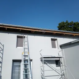 Szara elewacja budynku z nową rynną, widoczny fragment ocieplenia pod dachem, okna i rozstawione rusztowanie oraz drabina.