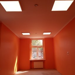 Kama-bud Usługi Ogolnobudowlane Kamil Tylkowski - Pomarańczowy pokój po remoncie, z białym grzejnikiem pod oknem i czterema kwadratowymi lampami sufitowymi.