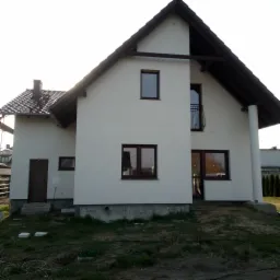 Dwukondygnacyjny dom jednorodzinny z białymi ścianami i brązowym dachem, widok z zewnątrz, z widocznymi oknami i drzwiami wejściowymi, teren wokół domu jeszcze niezagospodarowany.