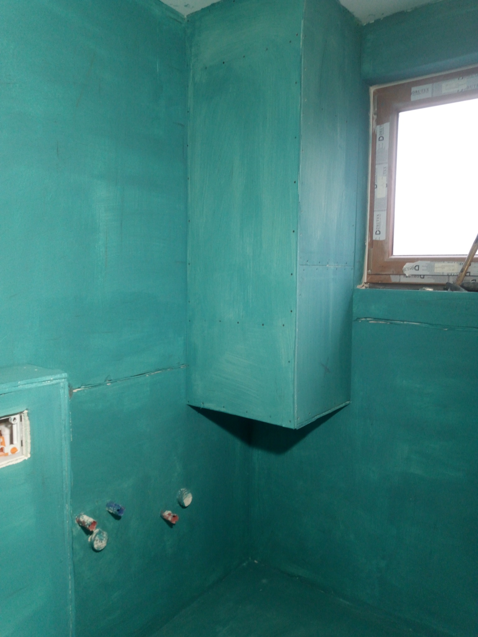 Przygotowana do dalszych prac łazienka, ściany i obudowa rur pokryte hydroizolacją w kolorze turkusowym, widoczne wystające rury instalacji wodnej i okno.