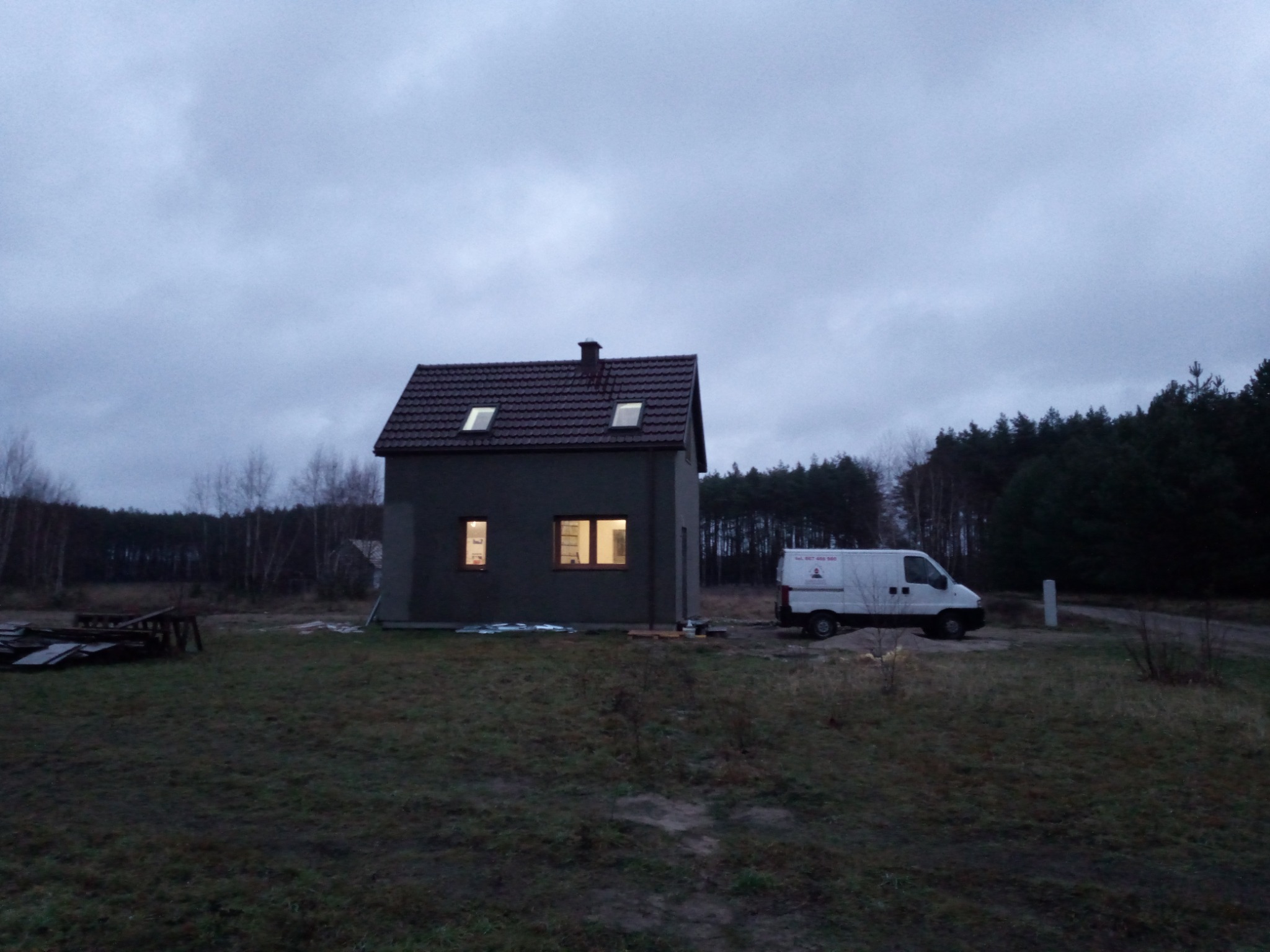 Niewielki, szary dom z ciemnoczerwonym dachem i oświetlonymi oknami, stojący na zewnątrz w pochmurny dzień, z zaparkowanym białym vanem z boku i lasem w tle.