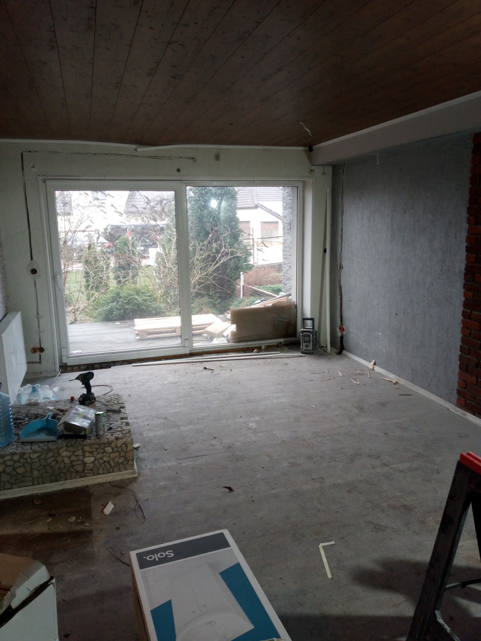 Pokój w trakcie prac wykończeniowych: surowy beton na podłodze, ściana z szarą fakturą i fragment ściany z cegły, drewniany sufit, okno balkonowe z widokiem na ogród, narzędzia i materiały...