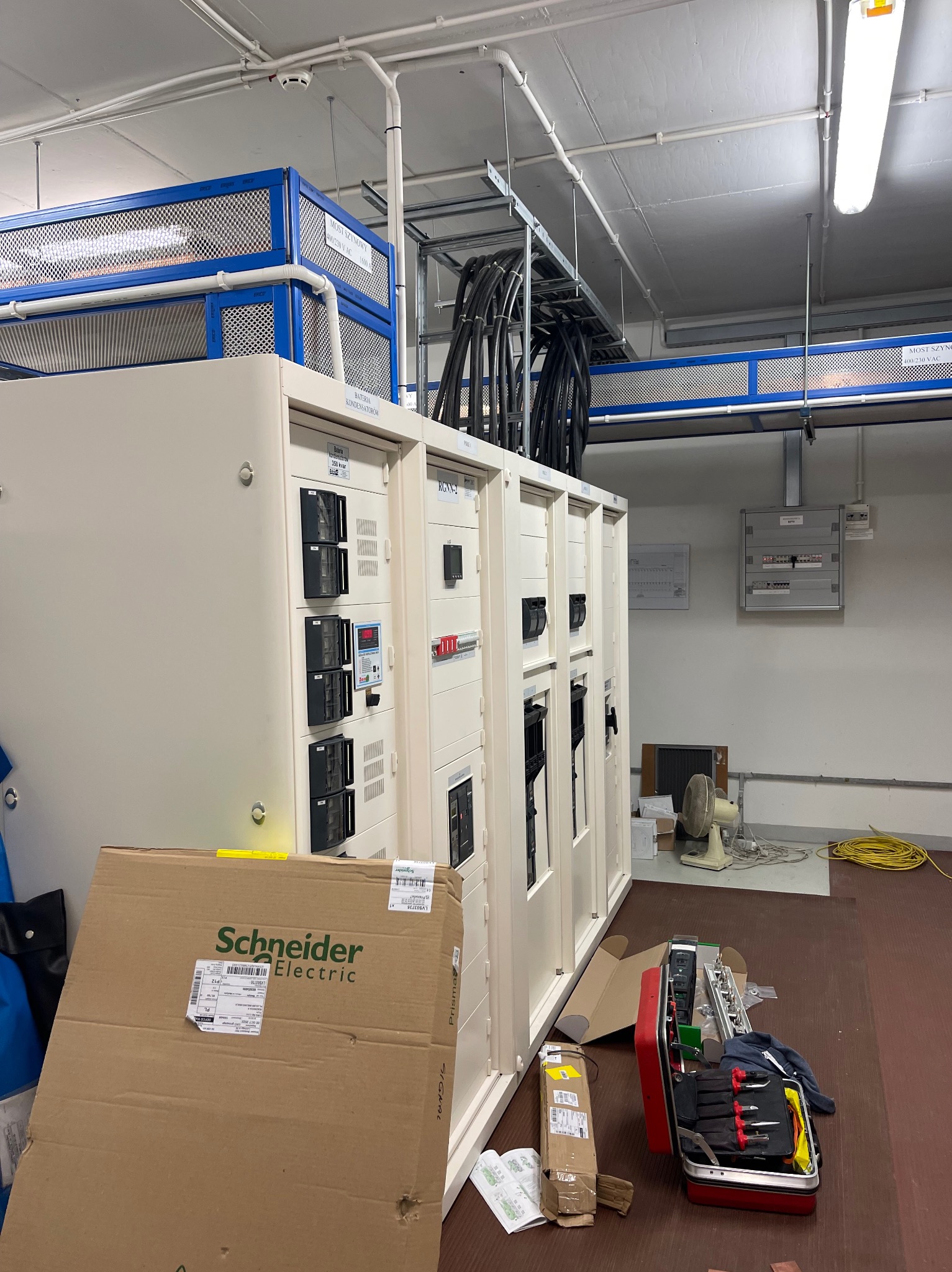 Rozdzielnica elektryczna w pomieszczeniu technicznym, widoczne kable, skrzynka narzędziowa i karton Schneider Electric. Elementy instalacji elektrycznej w tle.