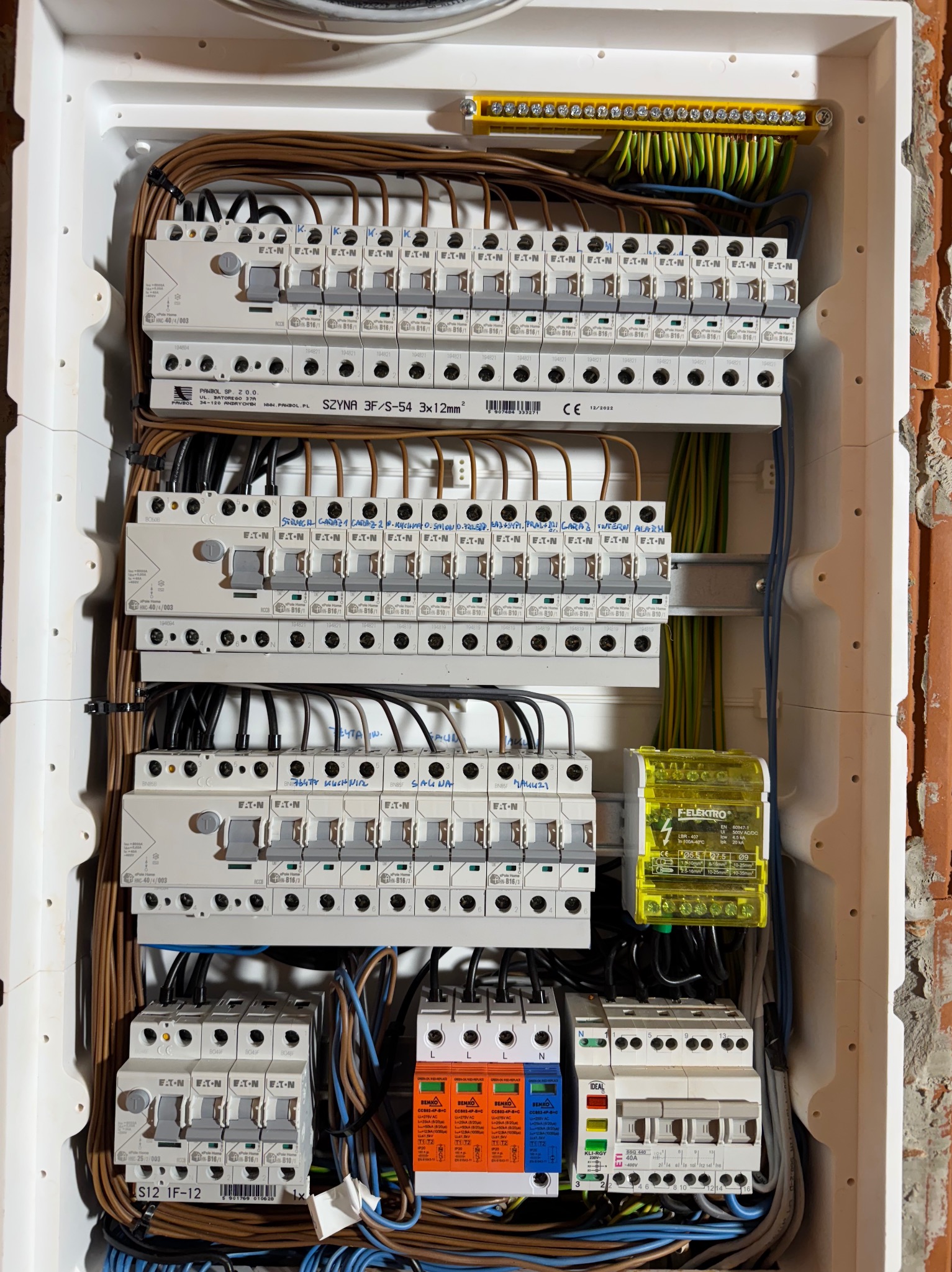 Rozdzielnia elektryczna z zabezpieczeniami, widoczne bezpieczniki EATON, przewody i listwy zaciskowe. Instalacja w białej obudowie na tle ceglanej ściany.