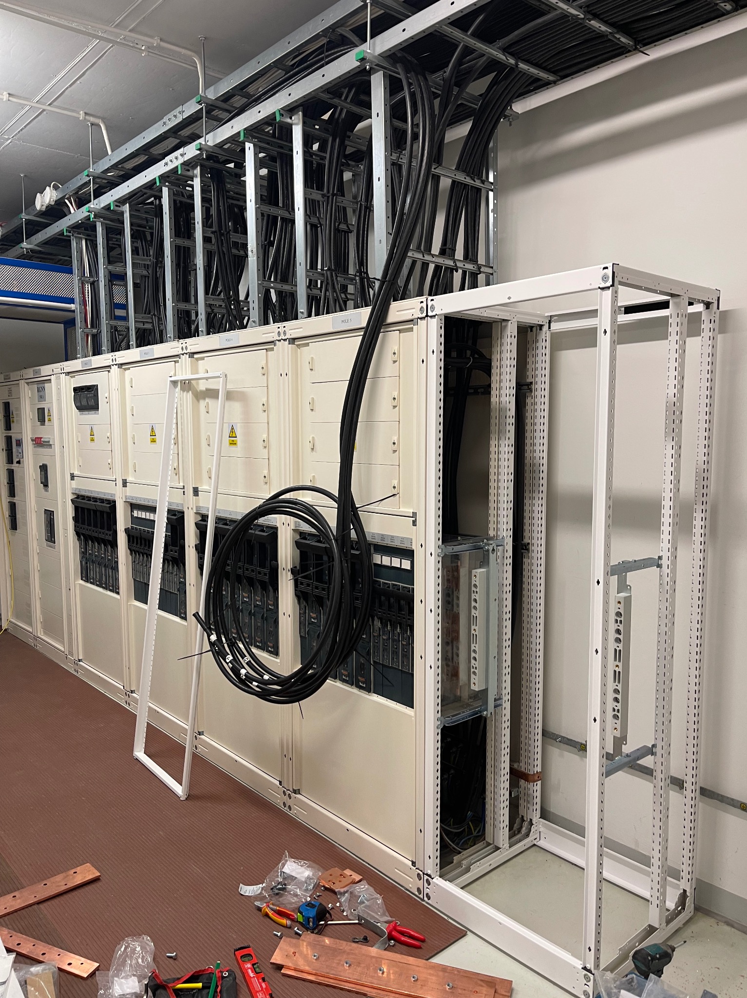 Rozbudowa rozdzielnicy elektrycznej: widoczne kable w korytkach kablowych, metalowa rama szafy i miedziane szyny. Etap prac instalacyjnych, narzędzia na podłodze.