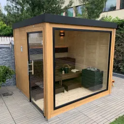 Drewniana kabina z przeszklonymi ścianami i czarnym dachem, wyposażona w piec na kamienie, drewniane ławki, oświetlenie i akcesoria do relaksu, umieszczona na tarasie w ogrodzie.